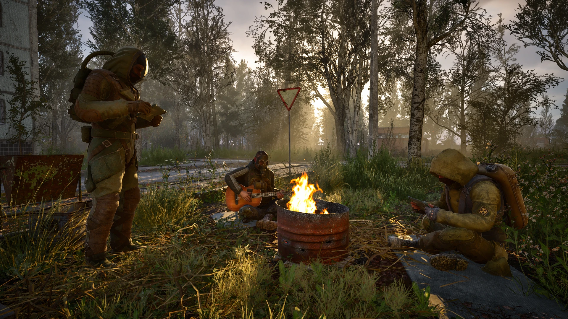 Three figures in hooded protective suits and gas masks gathered around a burning barrel in a grassy clearing; one stands reading, one sits playing a guitar, one crouches beside the fire.