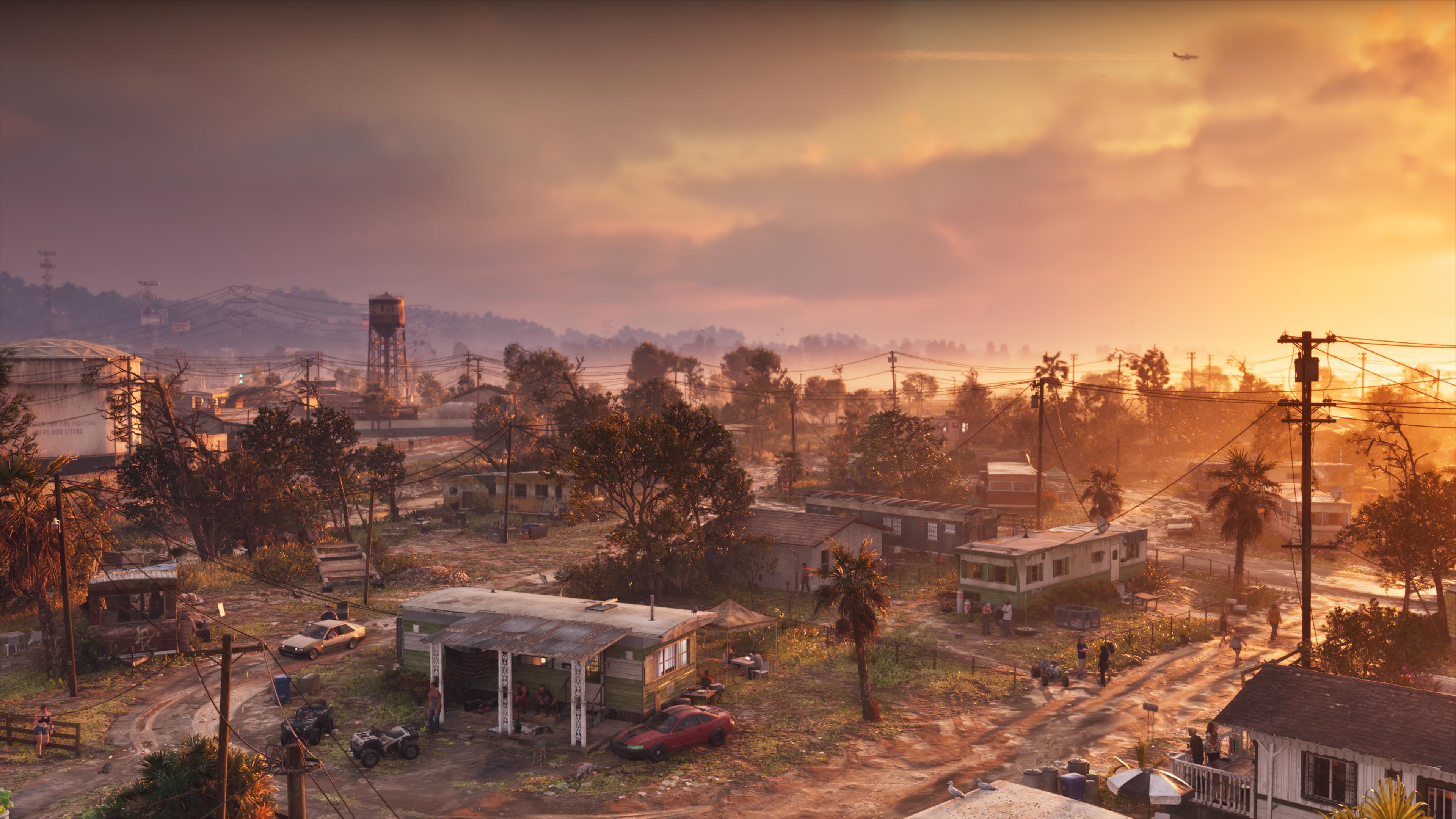 A small town of low houses, a water tower and palm trees seen from above under an orange sunset sky.